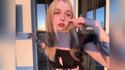 Blonde woman in a black ruffled top posing on a sunlit balcony during golden hour.