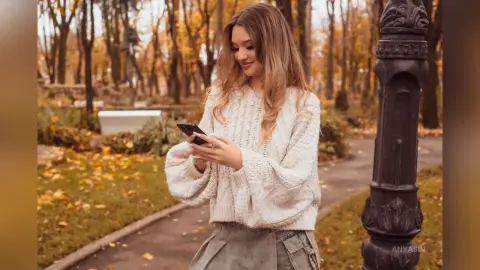 Young woman in a cozy white knit sweater using her smartphone in an autumn park setting.