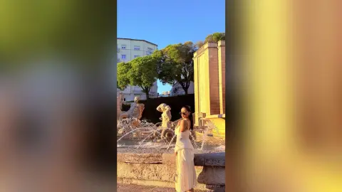 Stylish woman in a white summer dress standing by a stone fountain in a sunlit European city plaza.
