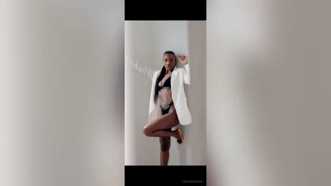 Confident Black woman in black lingerie and white blazer posing against a minimalist white wall.