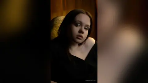 Moody portrait of a young woman in a black top sitting in a dimly lit room.