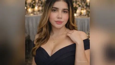 Beautiful woman in a navy blue off-the-shoulder dress and gold necklace posing in a soft, elegant setting.