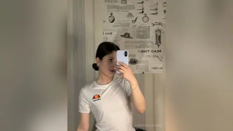 Young woman in a white graphic t-shirt taking a mirror selfie in a modern room with newspaper wallpaper.