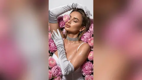 Glamorous woman in silver evening gloves and sparkling choker posing amidst pink peonies in a dreamy setting.