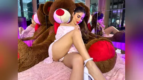ValeryRouge sits on a pink bed next to a large teddy bear, wearing a white lace chemise that exposes her thighs and chest.