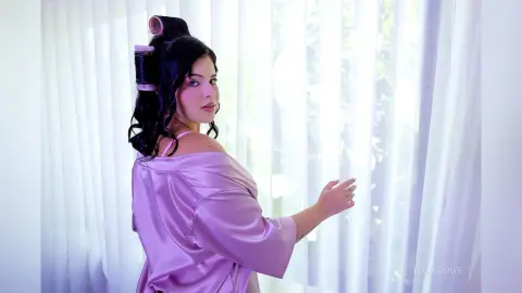 Young woman in a pink satin robe with hair rollers getting ready by a bright window in a soft, morning mood.