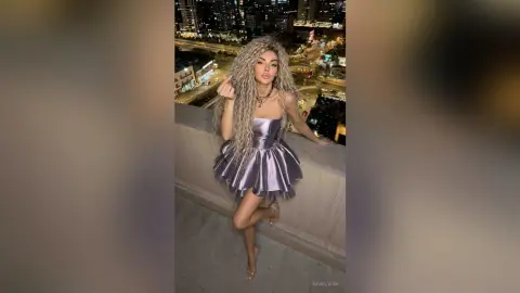 Glamorous woman in a metallic purple mini dress posing on a city balcony at night.