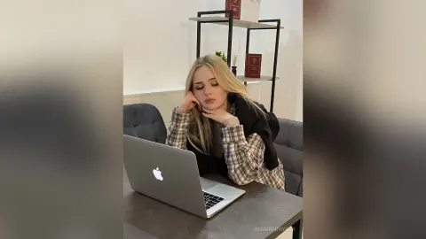 Blonde woman in a plaid shirt leaning on a desk with a laptop in a modern, minimalist home office setting.