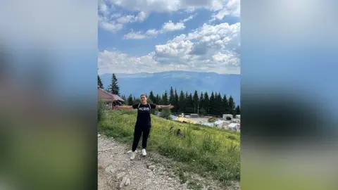 Young woman in black t-shirt and leggings smiling on a scenic mountain trail under a cloudy blue sky.