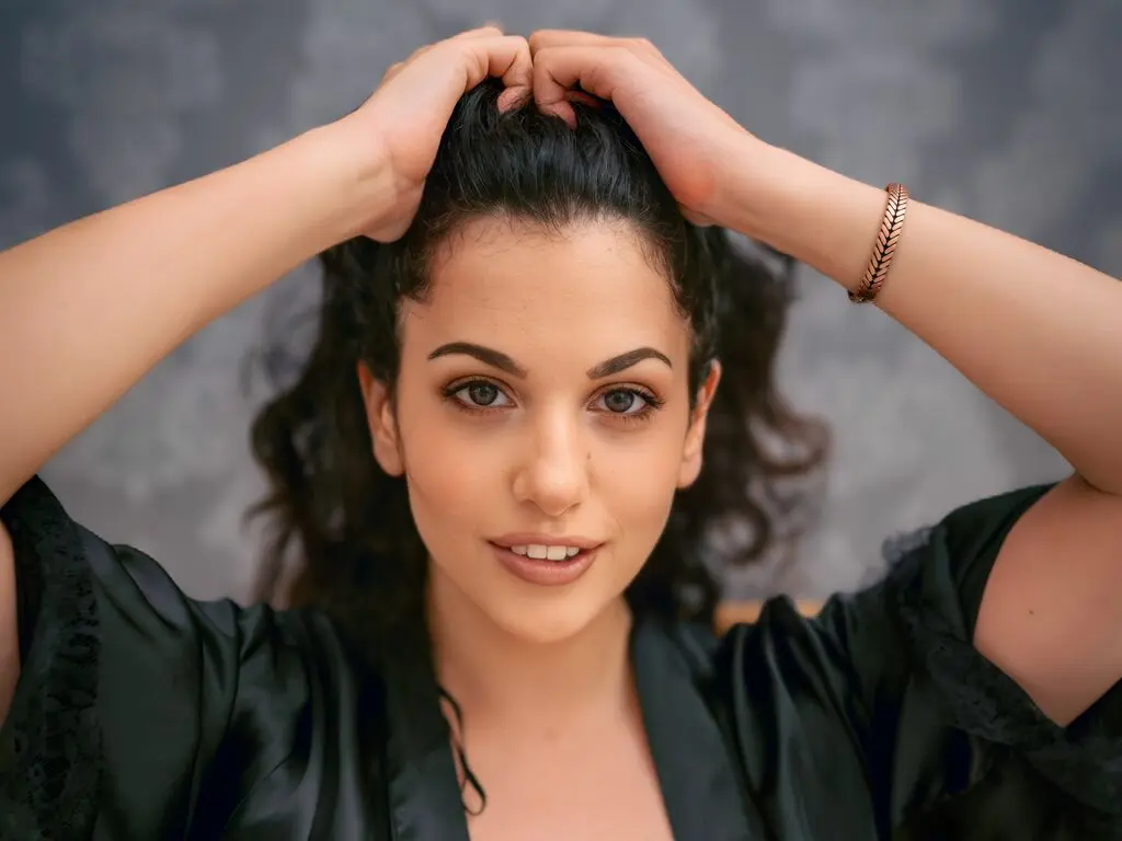 OrriHart poses with her hands in her dark hair, wearing a black robe against a grey background.