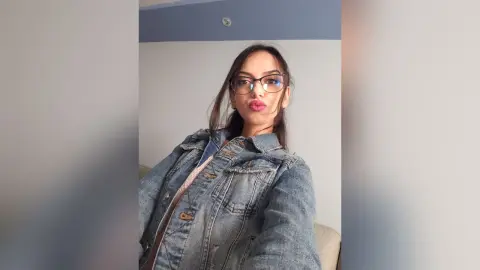 Young woman with glasses wearing a denim jacket posing in a bright indoor setting.