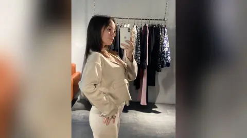 Young woman in a beige blazer taking a mirror selfie in a modern clothing boutique.