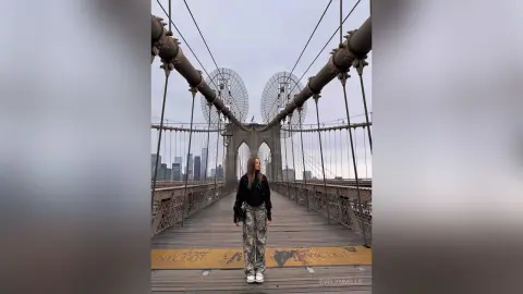 EvelynMills stands on the wooden planks of the Brooklyn Bridge wearing a black long-sleeve top and patterned grey trousers.