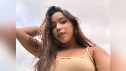 Young woman with long brown hair wearing a beige ribbed tank top against a cloudy sky background.