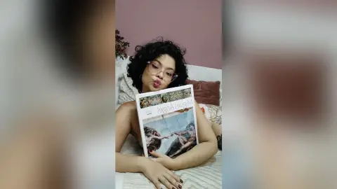 Curly-haired woman with glasses holding a Miguel Angel art book while relaxing on a bed in a cozy room.