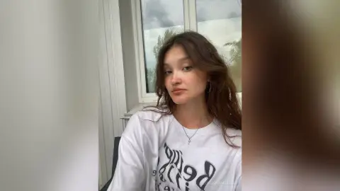 Young woman in a white graphic t-shirt sitting by a bright window in a relaxed, natural mood.
