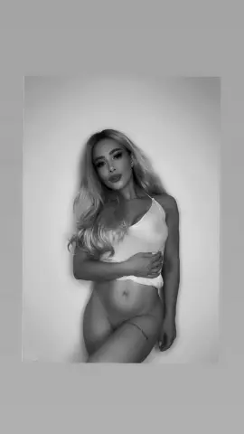 Blonde woman in white camisole posing against a plain wall in a moody black and white portrait.