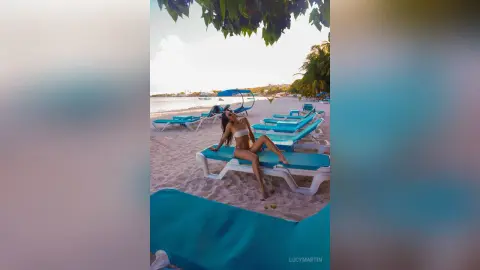 LucyMartin reclines on a white lounge chair on a sandy beach wearing a white bikini.