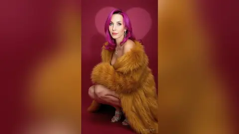 Elegant woman with pink hair in a luxurious mustard faux fur coat posing against a deep red background.