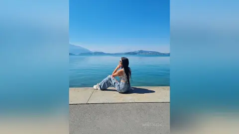 Young woman in denim jeans and a white crop top sitting on a pier overlooking a calm blue ocean under a clear sky.