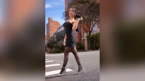 Stylish woman in a black cocktail dress and silver heels posing on a city street against urban buildings.