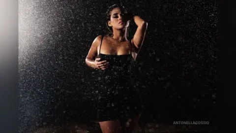 Moody portrait of a woman in a black tank top standing under heavy rain against a dark background.