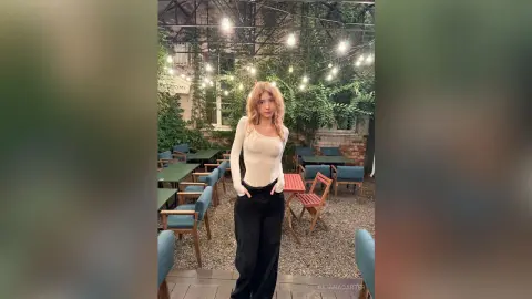 Young woman in a white long-sleeve top and black trousers standing in a cozy outdoor cafe at night.