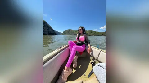 Woman in pink yoga outfit sitting on a boat on a sunny lake surrounded by mountains.