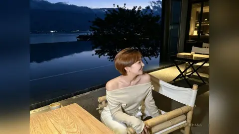 Smiling woman in an off-the-shoulder cream top relaxing by a luxury infinity pool at twilight.