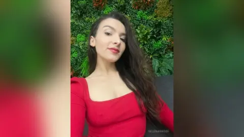 Young woman in a red dress smiling against a lush green botanical wall background.