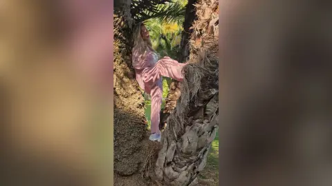 Stylish woman in a pink satin lounge set leaning against a textured palm tree in a sunny tropical garden.