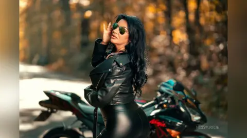 MissyJolie poses on a black motorcycle wearing a black leather jacket and tight leather pants against a blurred forest background.