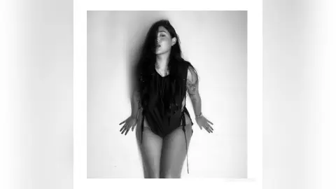 LaurenKaterine poses in a black bodysuit against a plain wall, showing her long dark hair and arm tattoos.