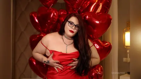 Curvy woman in a red satin slip dress posing against red heart balloons for a romantic Valentine's Day mood.
