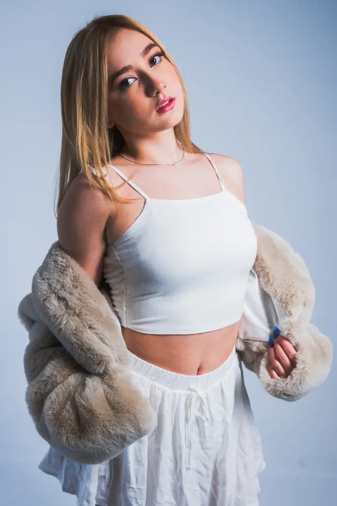 Sophiie_Collins — Sophiie_Collins poses in a white tank top and white skirt, wearing a tan faux fur coat over her shoulders.