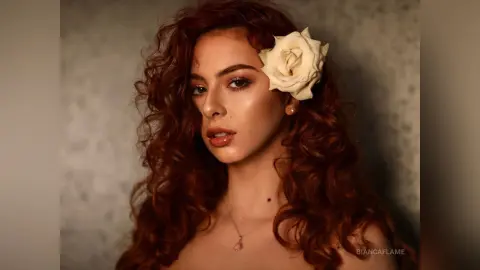 Portrait of a woman with curly red hair and a white rose accessory in a moody, cinematic studio setting.