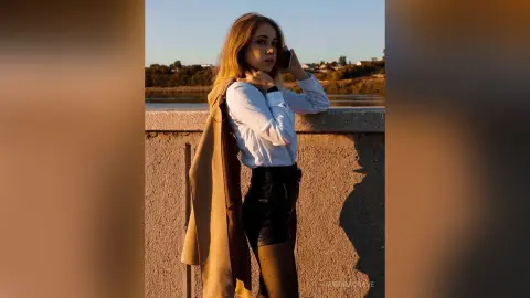 Young woman in a white blouse and black leather skirt posing by a riverside at sunset.