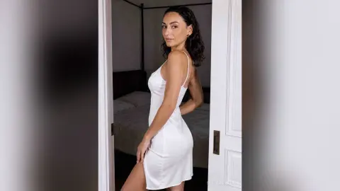 Elegant woman in a white silk slip dress posing in a minimalist bedroom setting.