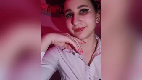 ChristyWeb wearing a light pink button-up shirt, looking at the camera with her hand resting near her chin against a red background.
