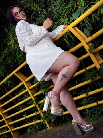 Woman in white lace dress and gold platform heels posing by a yellow railing in a lush green garden.