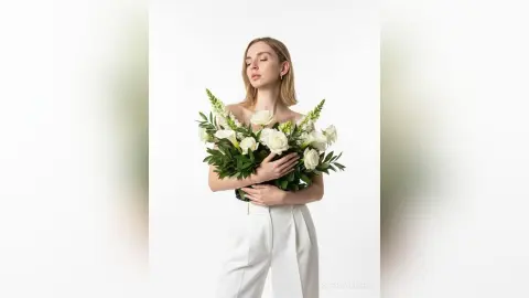 Blonde woman in white trousers holding a large bouquet of white flowers against a minimalist white background.