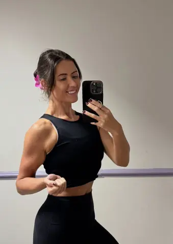 Fit woman in black athletic wear taking a mirror selfie in a bright dance studio with a pink flower in her hair.