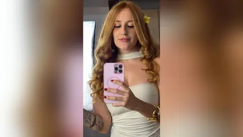 Redheaded woman in a white halter dress taking a mirror selfie in a bright room.