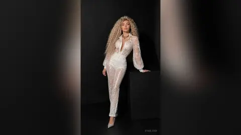 Elegant woman in a sparkling white lace jumpsuit posing against a dark studio background.