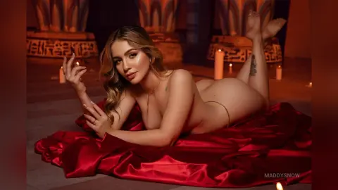Blonde woman in gold jewelry lying on red satin fabric in a moody, candlelit ancient Egyptian temple setting.