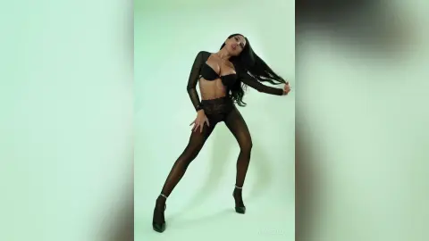 NicoleBella poses in black lace lingerie and sheer black tights against a plain light green background.