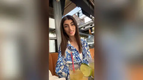Smiling young woman in a blue floral print shirt enjoying a refreshing lime cocktail at an outdoor cafe.