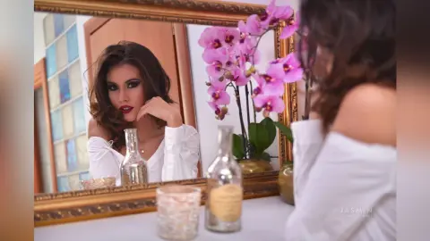 SandraMidnight poses in front of a gold mirror, wearing a white off-the-shoulder top with her dark hair styled in waves.
