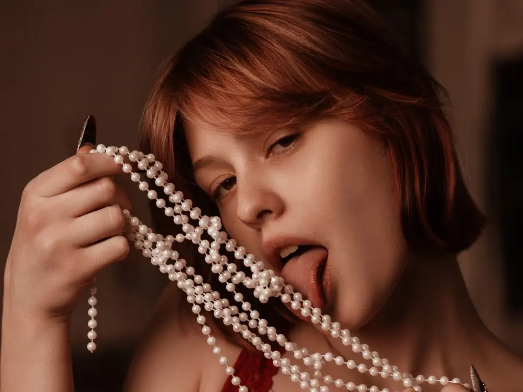 Young woman with red hair wearing a red outfit holding long white pearl necklaces in a moody indoor setting.