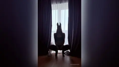 Silhouette of a woman in lingerie posing by a bright window in a moody, dark room.